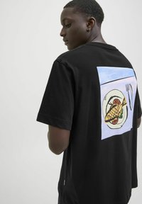 Sort bomulds t-shirt med et farverigt grafisk motiv på bagsiden af en fiskeanretning med en gaffel, sat mod en lyseblå baggrund.