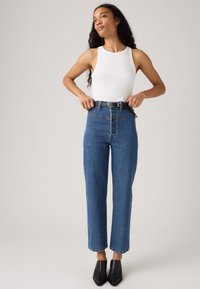 Donna che regola una cintura nera su jeans blu a vita alta, indossando un top senza maniche bianco e scarpe nere a punta, in piedi su uno sfondo neutro.