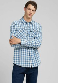 Camicia a maniche lunghe a quadri blu e bianchi con colletto abbottonato, due tasche sul petto e orlo curvo, indossata sopra jeans scuri.