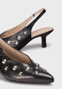 Zapatos de tacón negro de cuero con tiras al tobillo y punta afilada, decorados con tachuelas metálicas plateadas y un diseño elegante y minimalista.