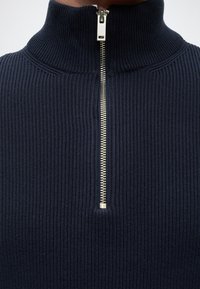 Primo piano di un maglione a coste blu navy con collo alto e mezza cerniera argentata al centro davanti.