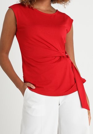 Vrouw die een mouwloze rode top met een zijstrikdetail draagt, gecombineerd met een hoge taille witte broek, hand in de zak.