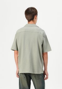 Camisa corta de manga corta texturizada en verde con cuello, que presenta un patrón sutil. La camisa se combina con jeans oscuros de corte holgado.