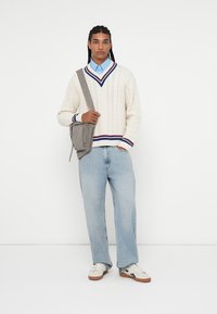 Pleteni krem pulover z mornarsko modrimi in burgundskimi poudarki, svetlo modra srajca s ovratnikom, svetle jeans hlače, siva torba čez ramo in bele superge.