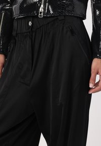 Pantalons noirs légers avec une taille élastique, dotés de poches latérales zippées et d'une texture lisse et mate. Accentué par une veste brillante ornée de sequins.