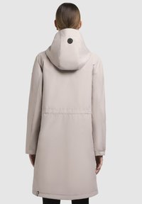 Doudoune beige clair avec capuche, présentant une taille cintrée, des lignes épurées et un logo noir rond au dos. Tissu lisse et texturé.