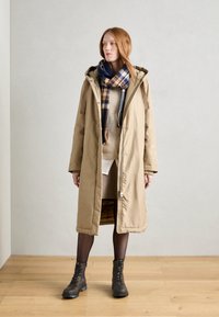 Beige dlouhý parka kabát s kapucí, předním zipovým zapínáním a bočními kapsami. Nositý přes pletený svetr a kostkovaný šál. Hnědé šněrovací boty.