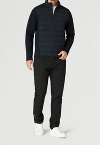 Chaqueta azul marino con parte delantera acolchada, mangas texturizadas, cierre completo de cremallera; combinada con pantalones negros y zapatillas blancas, diseño minimalista.