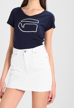 Kobieta nosząca granatowy t-shirt z białym abstrakcyjnym wzorem oraz białą, wysoką spódniczkę mini z denimu z postrzępionym brzegiem.