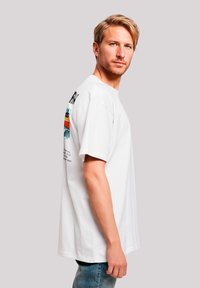 Weißes Baumwoll-T-Shirt mit kurzen Ärmeln. Mit buntem Grafikdruck auf dem Rücken, rundem Ausschnitt und lässiger Passform. Hellblaue Jeans.