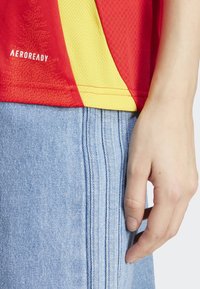Červené a žluté sportovní tričko s síťovanou texturou a logem AEROREADY, v kombinaci s světle modrými plisovanými džíny. Ruka odpočívající na šortkách.