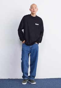 Camiseta preta de manga longa com logótipo branco, combinada com jeans azuis largos e ténis verdes, de pé contra um fundo simples.