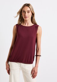 Mujer con cabello largo y rubio usa una blusa sin mangas de color burdeos, pendientes de aro, una pulsera oscura y pantalones color blanco roto, todo ello contra un fondo blanco liso.