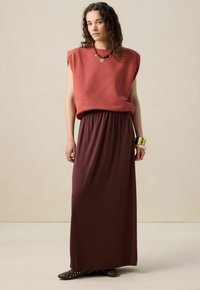 Haut sans manches rouge avec épaules tombantes, associé à une longue jupe fluide marron foncé. Les accessoires comprennent des bracelets en or et des chaussures décoratives.