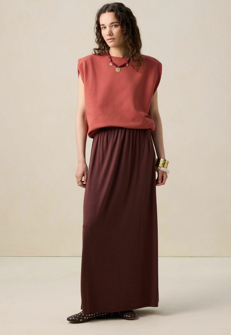 Haut sans manches rouge avec épaules tombantes, associé à une longue jupe fluide marron foncé. Les accessoires comprennent des bracelets en or et des chaussures décoratives.