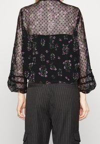 Blouse noire transparente avec motif floral rose et poignets en dentelle, portée avec un pantalon noir à rayures, vue de dos.