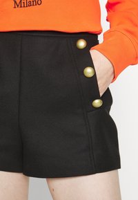 Main dans la poche d'un short noir taille haute avec trois grands boutons dorés, associée à un haut orange à manches longues portant l'inscription "Milano".