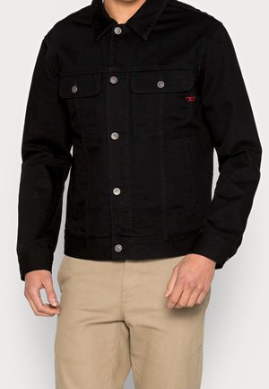 Homme portant une veste en jean noire boutonnée avec des poches poitrine et un pantalon beige, debout devant un fond uni.