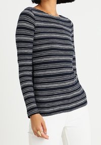 Haut de couleur marine à manches longues avec des rayures blanches horizontales, col rond, texture douce et coupe décontractée, associé à un pantalon blanc.