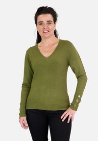 Suéter verde oliva de punto con escote en V, dobladillo acanalado y tres botones decorativos en cada puño. Combinado con pantalones negros.