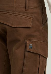 Bruine corduroy cargo broek met meerdere zakken, contrasterende stiksels en een metalen merkteken. Textiel met een stevige structuur.