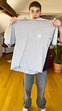 T-shirt cinza de manga curta feito de algodão, com um bolso e uma pequena etiqueta na frente. O modelo exibe a camisa com umas calças de ganga casuais.