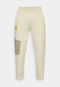 Κρεμ φόρμες με κίτρινο λογότυπο και λεπτομέρειες. Διαθέτουν ελαστική μέση, πλαϊνή τσέπη με φερμουάρ και σταμπωτές επιφάνειες σε αντίθετο χρώμα.