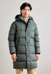 Puffer-takki tummanvihreästä kankaasta, jossa on huppu, vetoketjulliset taskut, napitus ja tikattu pinta. Pitkä, kapea malli.