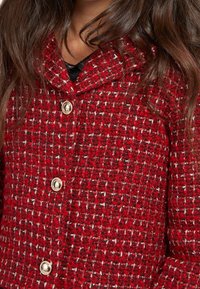 Manteau en tweed rouge avec un tissage texturé, présentant un col et des boutons dorés à l'avant. Vue rapprochée du détail du tissu.