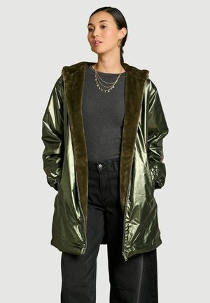 Vrouw die een glanzende olijfgroene lange jas met bontvoering draagt over een donkergrijze top en zwarte hoge taille broek, handen in de zakken, kijkt naar rechts.