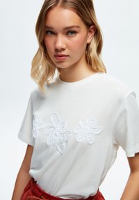 Camiseta de algodón blanca con tres diseños de flores bordadas en blanco. Cuello redondo, mangas cortas, textura suave y corte relajado.