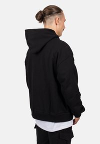Sudadera negra con un ajuste holgado, que cuenta con una gran capucha y puños de canalé, mostrada desde la espalda de una persona que lleva una prenda blanca debajo.