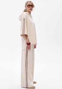 Vrouw in een beige capuchonloze top en wijde broek met donkere streepdetails op de mouwen en broekspijpen, die grote zonnebril en oorringen draagt.