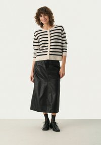 Gestreepte cardigan in crême en zwart, met knopen aan de voorkant, zachte textuur. Zwarte leren rok tot halverwege, aansluitend ontwerp, subtiele zijnaaddetails. Zwarte schoenen.