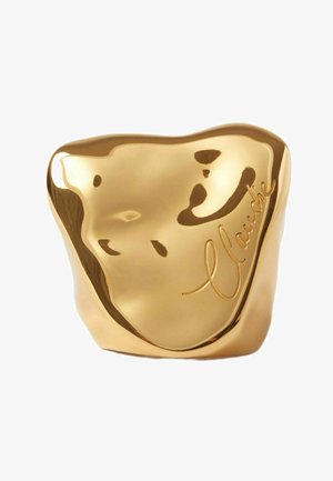 Scultura astratta color oro con superficie liscia e riflettente; presenta un testo inciso "Claude" e un semplice design di volto stilizzato.