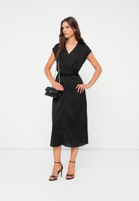 Black wrap dress with short cap sleeves, knee-length hem, and smooth texture. Paired with a small black handbag and strappy black heels.