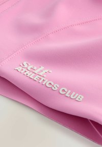 Tessuto atletico rosa liscio con finitura strutturata; presenta in evidenza il testo bianco "self. ATHLETICS CLUB". Sono visibili le cuciture.