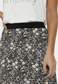 Jupe noire fleurie avec marguerites blanches et touches roses. Caractérisée par une ceinture élastique noire texturée. Tissu lisse et design plissé.