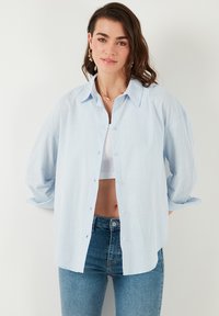 Camicia azzurra chiaro con bottoni realizzata in tessuto testurizzato, caratterizzata da un colletto, maniche oversized e una vestibilità rilassata. Indossata sopra un top bianco.