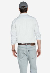 Camisa blanca a rayas con mangas largas, gorra gris, jeans azul claro y cinturón trenzado marrón, vista desde la espalda.