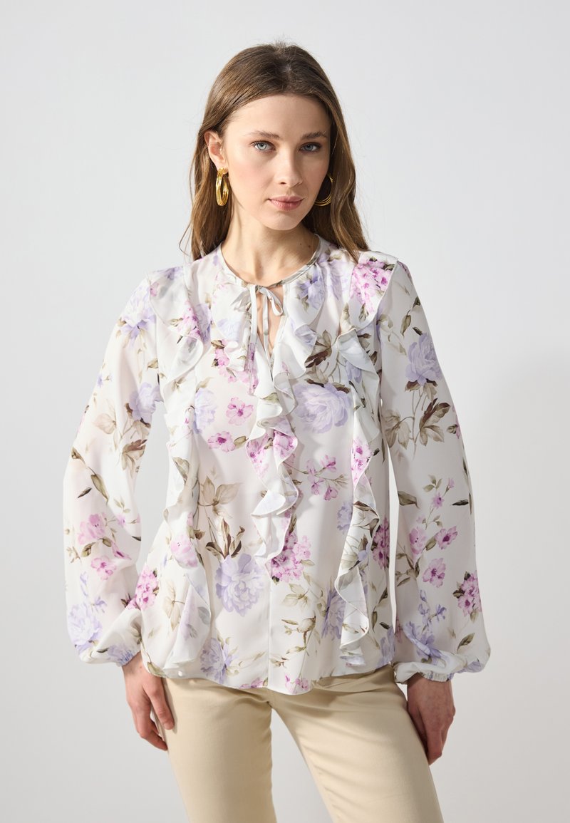 Hvit bluse med blomster og rysjer, med lilla og rosa blomster og knyting i halsen. Lange ermer med elastiske mansjetter. Glatt stoff.