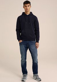 Šviesiai mėlynos spalvos vyrų gobtuvu su priekine kišene, derinamos su mėlynais džinsais. Modelis nešioja pilkas sportines batus su juodais akcentais.