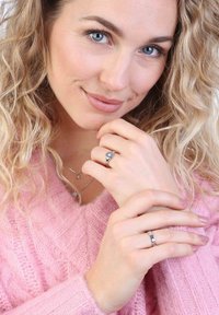 Zilveren ringen met een centrale edelsteen, gedragen op de vingers. Het model heeft golvend blond haar en draagt een roze gebreide trui.