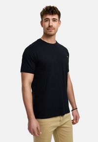 Sort kortærmet t-shirt lavet af bomuld, med en lomme og diskret farveaccent, båret med beige bukser. Glat tekstur.