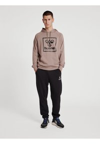 Brun huvtröja med en svart logotyp, tillsammans med svarta sweatpants som har ett vitt emblem, och svarta sneakers med blå accenter.