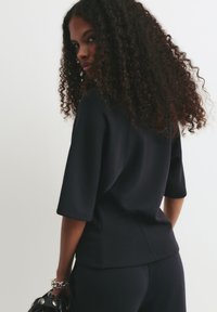 Femme avec de longs cheveux bouclés portant un haut noir à manches courtes et un pantalon, tenant un sac noir avec des clous en argent, regardant par-dessus son épaule.