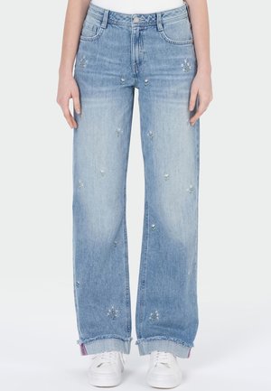 Hellblaue Jeans mit weitem Bein, kleinen weißen Blumenstickereien und ausgefransten Säumen, getragen mit weißen Turnschuhen.
