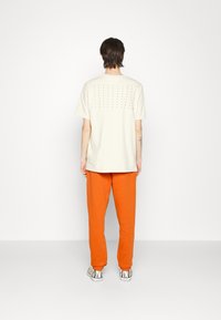 Ljust beige t-shirt med perforerat prickmönster på ryggen, kombinerat med klara orange byxor och mönstrade skor.