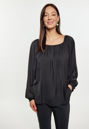 Frau mit langen braunen Haaren, die eine lockere schwarze Bluse mit langen Ärmeln und Perlenohrringen trägt, steht vor einem schlichten hellen Hintergrund.