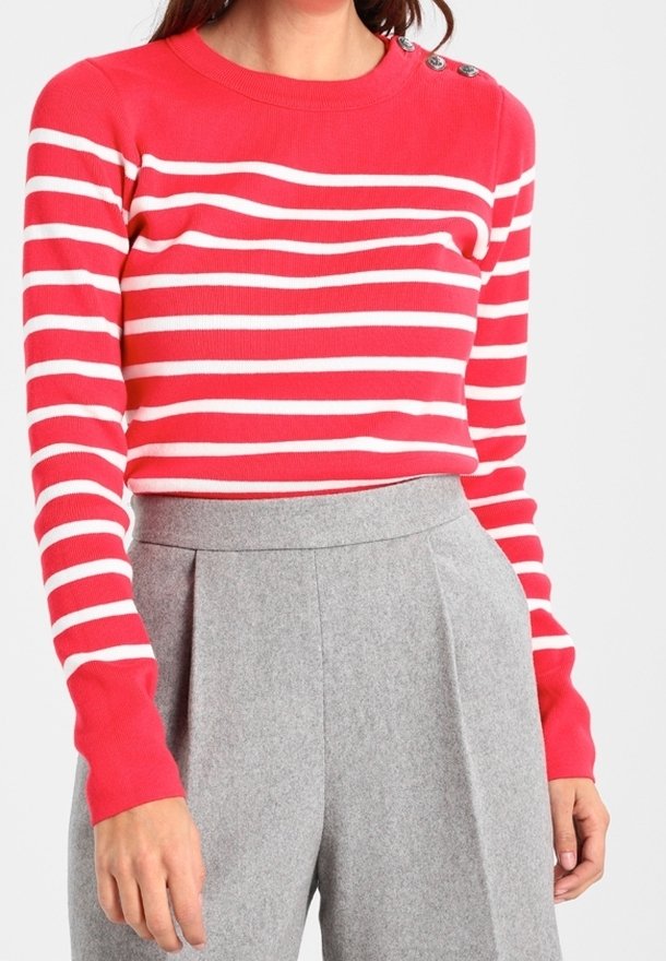Femme portant un pull rayé horizontal rouge et blanc avec des boutons aux épaules et un pantalon plissé gris taille haute.